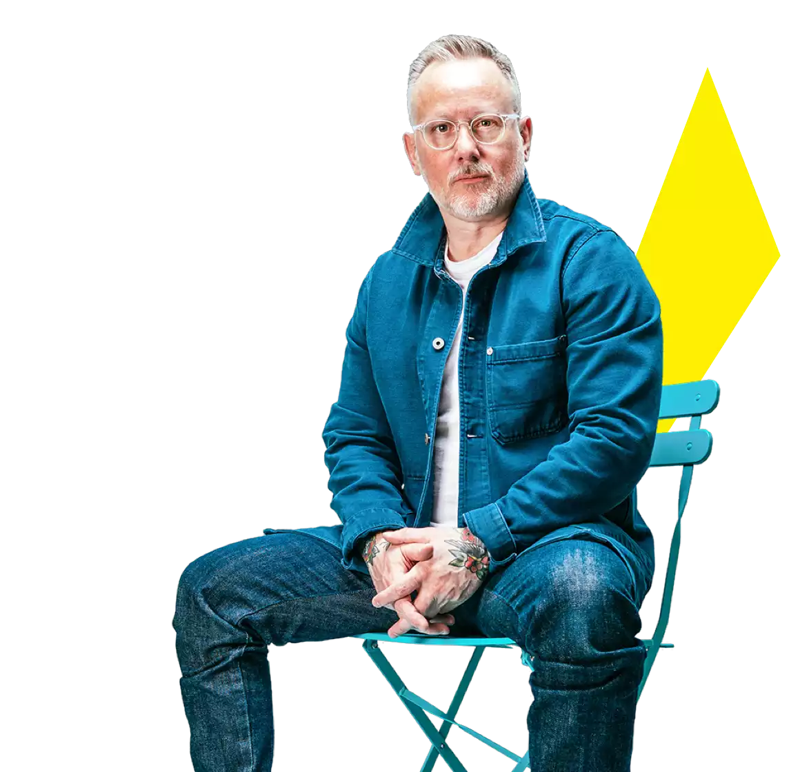 a man with glasses sitting down on a blue chair, wearing a jeans and jean jacket, and looking straightahead