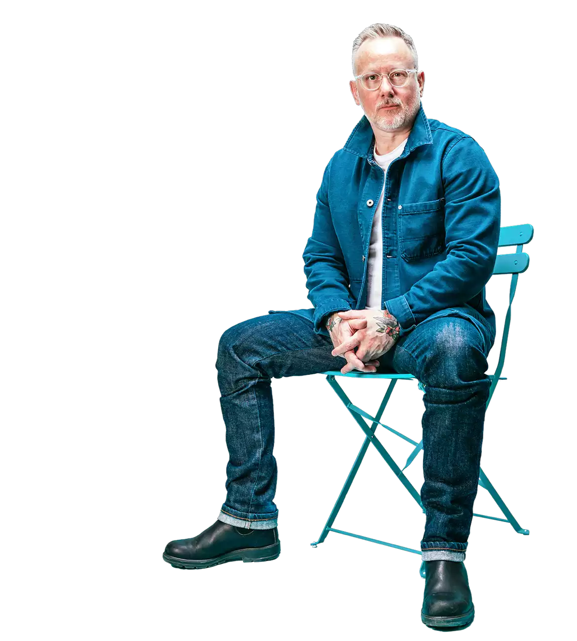 a man with glasses sitting down on a blue chair, wearing a jeans and jean jacket, and looking straightahead